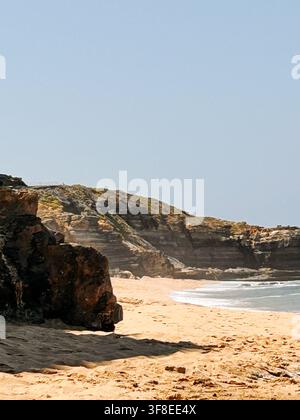 Plage de sable paisible bordée de falaises rocheuses stratifiées dramatiques, avec des vagues océaniques douces et un gros rocher au premier plan Banque D'Images