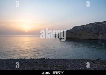 Couchant doré sur une mer calme et une côte montagneuse escarpée en Oman Banque D'Images