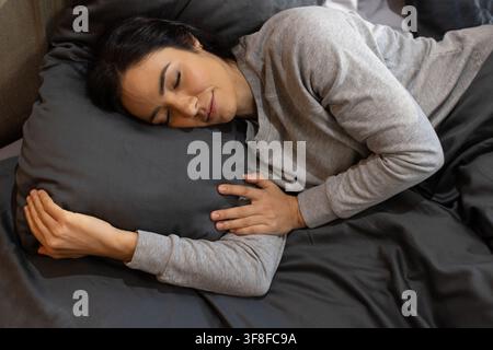Femme allongée sur le lit portant un haut à manches longues gris clair, reposant sa tête sur un oreiller gris foncé. Sommeil, couette, tête de lit, coussin, chambre, atmosphérique, confortable Banque D'Images