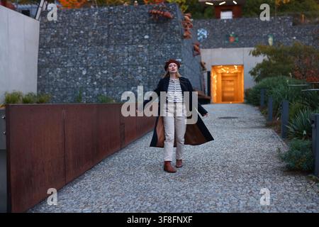 Femme élégante dans une tenue inspirée du style français posant à l'extérieur, portrait de style de vie avec architecture moderne et atmosphère automnale. Banque D'Images