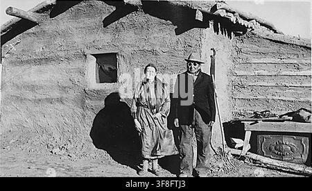 Un couple indien debout devant une cabane en rondins enduite de boue, photographiée en 1939, dans le cadre des archives du Bureau of Indian Affairs. Banque D'Images