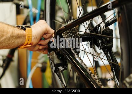 Mécanicien cycliste masculin ajustant la transmission arrière d'un vélo de route pendant l'entretien. Un gros plan montre ses mains travaillant près du cassette, de la chaîne et Banque D'Images