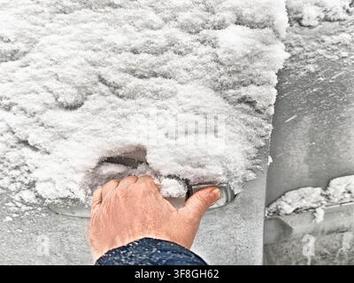 Vue rapprochée d'une personne luttant pour déverrouiller une poignée de porte de voiture recouverte de glace et de neige par temps froid d'hiver. Banque D'Images