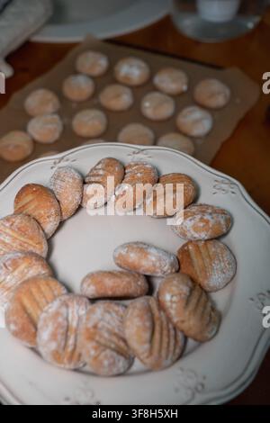 Cookies aux amandes fraîchement cuits saupoudrés de sucre glace refroidissant sur du papier sulfurisé avec une autre fournée servie sur une assiette au premier plan. Pâtisserie maison, dessert, r Banque D'Images