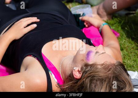 FEMME AVEC UN VISAGE EN Poudre SCINTILLANTE COUCHANT SUR L'HERBE, REPORTAGE, FESTIVAL GREEN MAN 2016, BRECON, WALES, ROYAUME-UNI, 2016 : Une femme avec du glitter violet sur la joue repose sur l'herbe les yeux fermés, portant un débardeur noir par-dessus un soutien-gorge rose. Photo : Rob Watkins. INFO : Le Festival Green Man a débuté en 2003 au Pays de Galles. Il se tient chaque année dans les Brecon Beacons le troisième week-end d'août et célèbre la musique alternative, les arts, la littérature, la science et une culture familiale dans un magnifique cadre montagneux. Banque D'Images
