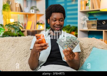 Jeune Afro-Américain sur le canapé à la maison compte de l'argent, des dollars en espèces et des chèques sur son smartphone après une victoire à un jeu en ligne dans le salon. Le type noir compare le montant et profite d’un succès aux paris sportifs ou au casino à l’intérieur. Banque D'Images