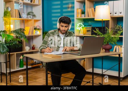 Homme indien assis à une table à domicile porte des écouteurs, écoute une leçon et prend des notes avec un sérieux concentré. Homme arabe assis à une table à domicile hoche la tête pendant un cours en ligne ou un appel client en utilisant un ordinateur portable pour une formation en ligne. Banque D'Images