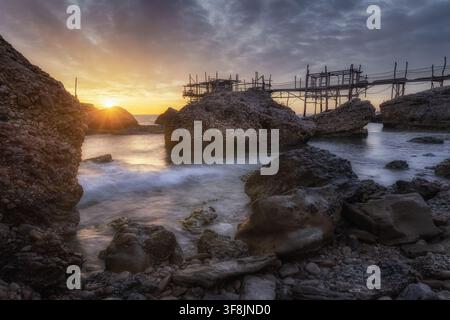 Vue du Trabocco Spezzacatena au coucher du soleil avec une côte rocheuse, des vagues et une lumière dorée à Rocca San Giovanni, province de Chieti, Italie. Banque D'Images