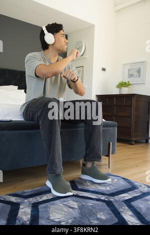 Homme asiatique assis au bord du lit portant des écouteurs blancs et vérifiant sa montre-bracelet près d'un smartphone Banque D'Images