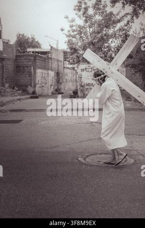 Une vue de rue en noir et blanc grand angle d'un homme déguisé en Jésus portant une grande croix en bois le long d'une route urbaine déserte. Banque D'Images