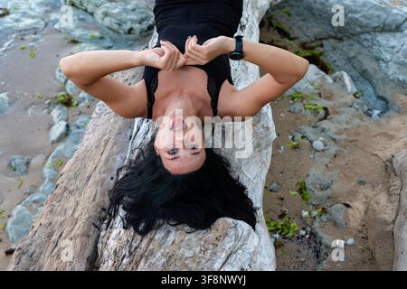 Femme souriante allongée à l'envers sur un bois flotté à la plage, créant une ambiance estivale joyeuse et détendue pour les concepts de style de vie et de bien-être. Banque D'Images