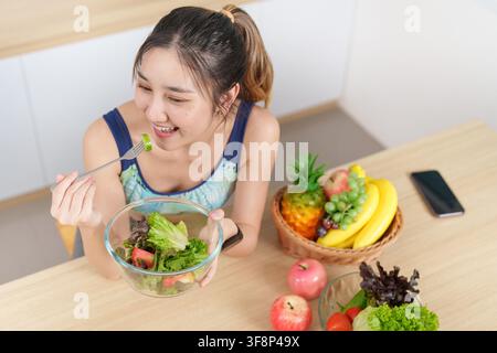 Femme asiatique heureuse mangeant un bol de salade fraîche dans la cuisine pour un mode de vie sain et le bien-être Banque D'Images
