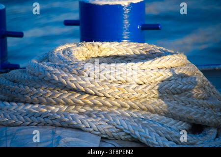 Corde d'amarrage épaisse enroulée reposant sur le pont d'un bateau à côté d'une bitte bleue, éclairée par une lumière chaude du soleil avec la mer en arrière-plan. Détail nautique en gros plan Banque D'Images