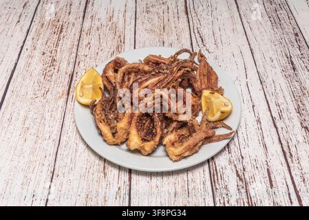 Anneaux et tentacules de calamars frits servis avec deux quartiers de citron sur une assiette blanche, fond en bois défraîchi. Banque D'Images