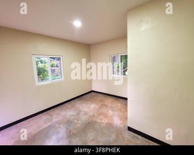 Une pièce vide et lumineuse avec un sol carrelé et deux fenêtres donnant sur des arbres verts, des murs peints en neutre et un éclairage encastré au plafond créant une ambiance calme et accueillante Banque D'Images
