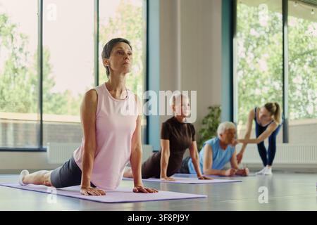 Femme caucasienne mûre pratiquant la pose du cobra lors d'un cours de yoga en groupe avec des participants âgés. L'image représente le vieillissement actif et le bien-être, idéale pour le marketing de la forme physique chez les seniors Banque D'Images