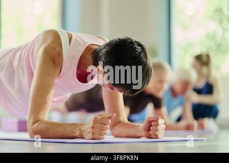 Femme caucasienne mûre effectuant un exercice de planche sur les avant-bras pendant un cours de fitness en groupe dans une salle de sport. L'image représente un mode de vie actif et convient au marketing du bien-être, prise de vue à faible angle Banque D'Images