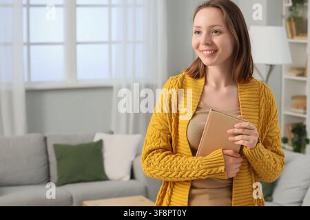Jeune femme souriante tenant un livre dans un salon confortable, concept d'éducation et d'apprentissage à domicile, le cadre décontracté suggère le confort et la détente w Banque D'Images