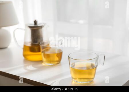 Théière en verre et deux tasses de thé aux herbes doré sur une table blanche avec une lumière naturelle et des ombres douces, une lumière naturelle douce entre par une fenêtre, projette Banque D'Images