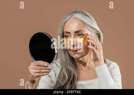 Femme caucasienne âgée appliquant des patchs pour les yeux devant un miroir à la maison, concept de soins de la peau Banque D'Images