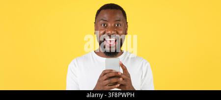 Homme afro-américain heureux tenant un smartphone et souriant, fond jaune Banque D'Images