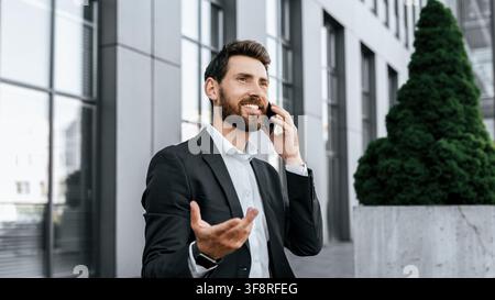 Homme d'affaires heureux parlant au smartphone à l'extérieur d'un bâtiment de bureaux moderne, concept commercial Banque D'Images