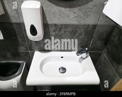 Vue rapprochée de sanitaires publics contemporains et propres. Mise au point sur le lavabo blanc, le robinet en métal brillant, le distributeur automatique de savon. Carrelage texturé sombre c Banque D'Images