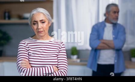 Femme caucasienne âgée inquiète aux bras croisés dans la cuisine, conflit relationnel émotionnel Banque D'Images