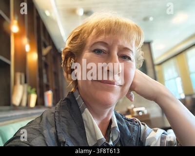 Une femme blonde d'âge moyen sourit pour un selfie tout en se détendant dans un café élégant, capturant l'essence de son expérience de voyage et de son style de vie. Banque D'Images