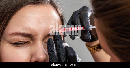 Injection de filler dans le front. Femme recevant une injection de filler botox anti-âge dans le front par une cosmétologue. Les mains de l'expert en beauté injectent Banque D'Images