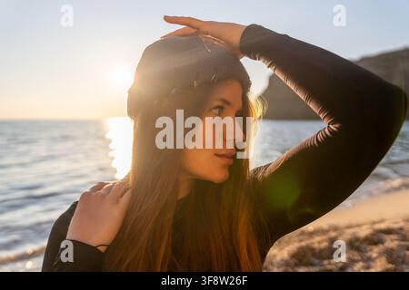 Femme, coucher de soleil, plage. Jeune voyageuse attirante portant un bonnet regarde la lumière dorée de l'océan à l'heure dorée pendant des vacances côtières. Banque D'Images