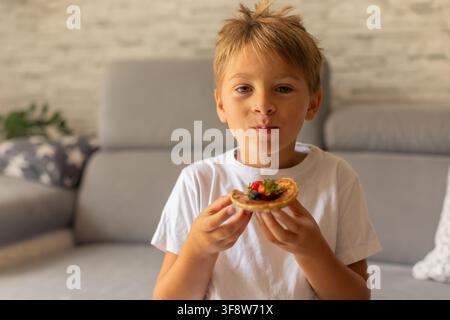 Petit enfant adorable, garçon, mangeant des pancakes américains avec des fraises, des cerises et des myrtilles, arrosés de sirop, chocolat et miel Banque D'Images