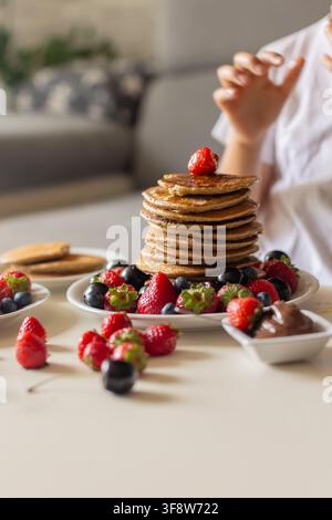 Petit enfant adorable, garçon, mangeant des pancakes américains avec des fraises, des cerises et des myrtilles, arrosés de sirop, chocolat et miel Banque D'Images