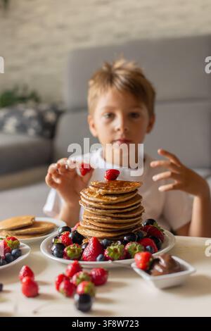 Petit enfant adorable, garçon, mangeant des pancakes américains avec des fraises, des cerises et des myrtilles, arrosés de sirop, chocolat et miel Banque D'Images