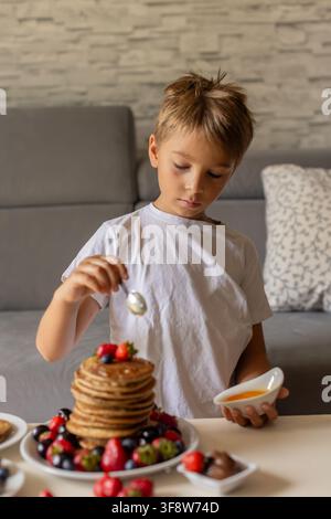 Petit enfant adorable, garçon, mangeant des pancakes américains avec des fraises, des cerises et des myrtilles, arrosés de sirop, chocolat et miel Banque D'Images