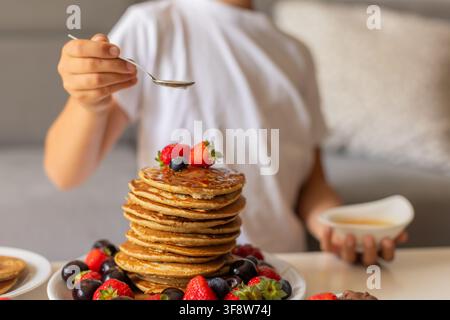 Petit enfant adorable, garçon, mangeant des pancakes américains avec des fraises, des cerises et des myrtilles, arrosés de sirop, chocolat et miel Banque D'Images