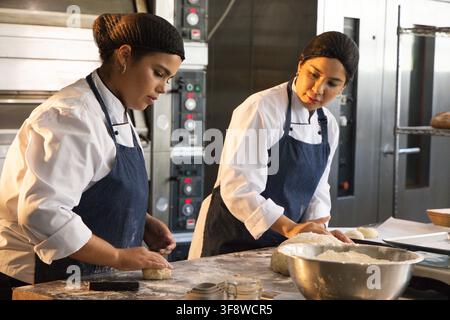 Deux chefs femmes façonnant la pâte sur une table de préparation farinée dans la cuisine de boulangerie, fours et portant des tabliers Banque D'Images