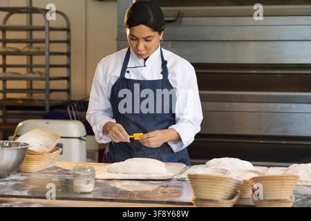Boulangère en veste de chef, tablier en jean travaillant au comptoir de boulangerie avec des bannetons façonnant trois pains Banque D'Images