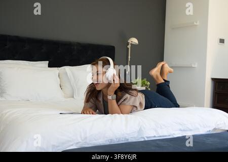 Femme afro-américaine adulte souriant allongée sur un lit blanc, tête de lit noire, tenant des écouteurs Banque D'Images