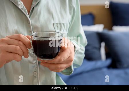 Femme en pyjama vert menthe tenant une tasse en verre transparent de café noir dans un cadre de chambre, la scène suggère une détente matinale et un confort au lit Banque D'Images