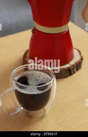 Café noir fraîchement infusé dans une tasse en verre à double paroi avec une cafetière rouge moka sur une table en bois, une cafetière rouge moka vive repose sur un dessous de verre en bois rustique Banque D'Images