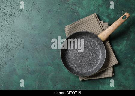 Poêle antiadhésive vide avec manche en bois sur une serviette de cuisine à carreaux et fond vert foncé, la poêle et la serviette sont positionnées sur un vert foncé Banque D'Images