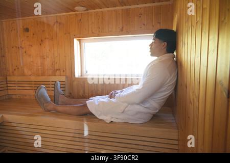 Homme asiatique assis sur un banc en bois dans un sauna portant une robe de chambre près d'une fenêtre rectangulaire, espace de copie Banque D'Images