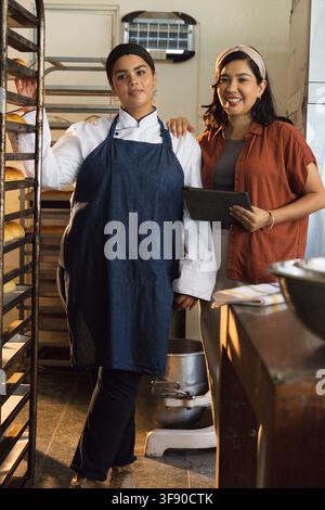 Diverses collègues féminines travaillant dans une boulangerie portant une blouse de chef au niveau d'une étagère avec des pains en utilisant une tablette Banque D'Images