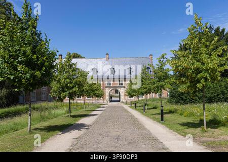 Un chemin pavé bordé d'arbres mène à la porte d'entrée de l'abbaye Abdij van Park à Louvain, en Belgique, encadré par des bâtiments historiques en brique, des pelouses vertes et un ciel d'été clair, évoquant un héritage paisible. Banque D'Images