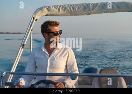Un homme confiant dirige un bateau à moteur au large d'Umag pendant l'heure dorée, portant des lunettes de soleil et une chemise blanche tout en profitant des loisirs d'été, de l'eau calme et des vues pittoresques sur la côte. Banque D'Images