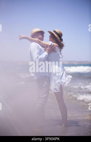 Romance debout s'appuyant l'un sur l'autre avec une stabilité confiante, s'assurant l'un l'autre, un couple portant des chapeaux s'embrasse sur une plage ensoleillée avec des vagues dans le Banque D'Images