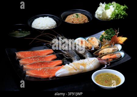 Un ensemble de fruits de mer et de saumon avec des crevettes, des calamars et du saumon tranché disposés sur un plateau noir avec une sauce de trempage. Banque D'Images