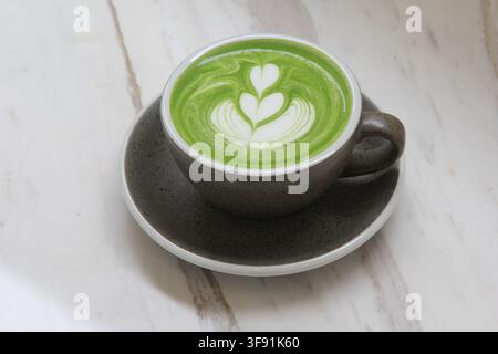 Une tasse chaude de latte au matcha vert avec un délicat art latte en forme de cœur servi sur une soucoupe sombre. Banque D'Images