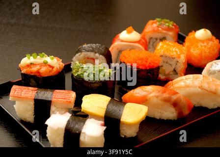 Vue en gros plan d'un plateau noir présentant une sélection de rouleaux de sushi japonais, des nigiri au saumon et des nigiri aux crevettes garnis de garnitures. Banque D'Images